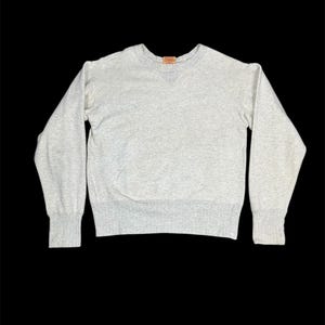 May include: A heather gray crew neck sweatshirt with long sleeves. The sweatshirt has ribbed cuffs and a ribbed hem. A small orange tag is visible at the neckline. The garment is displayed against a black background.