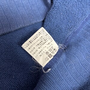 May include: Close-up of a blue garment with a white tag. The tag has Japanese text and care symbols. The garment is made of cotton.