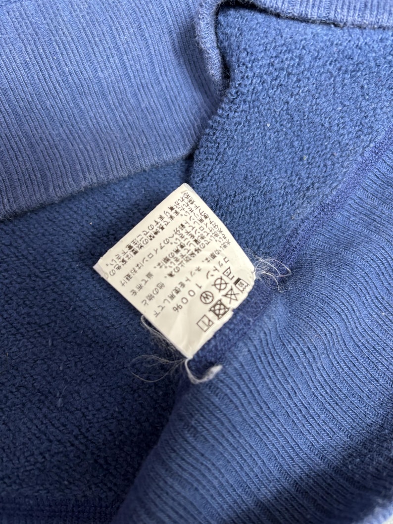 May include: Close-up of a blue garment with a white tag. The tag has Japanese text and care symbols. The garment is made of cotton.