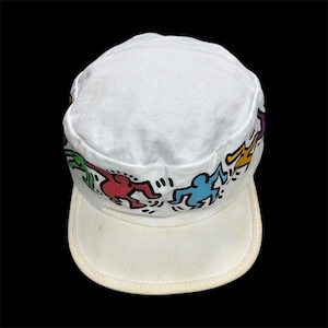 May include: White canvas cap featuring a colorful Keith Haring design. The cap has a flat brim and a structured crown. The design includes dancing figures in red, green, blue, yellow, and purple. A stylish accessory for casual wear.