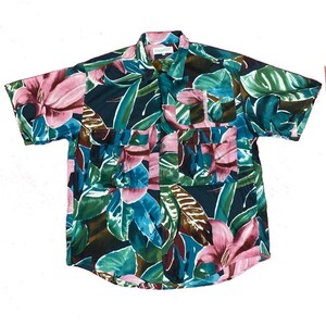 May include: Short-sleeved button-up shirt with a tropical floral pattern. The shirt features a dark teal background with large pink flowers, green leaves, and accents of blue and brown. It has two chest pockets and a classic collar.