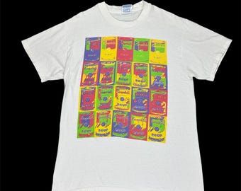 May include: White t-shirt with a pop art design of Campbell's soup cans. The graphic features multiple rows of soup cans in various colours, including red, yellow, blue, and green. The shirt has a classic crew neck style.