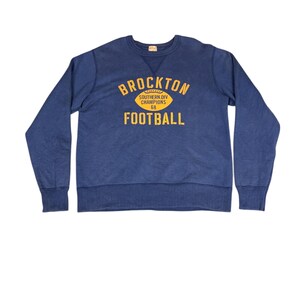 May include: A navy blue sweatshirt with the text "BROCKTON FOOTBALL" in yellow. The text is arched above a yellow football graphic. Below the graphic is the text "SOUTHERN DIV CHAMPIONS 68". The sweatshirt has ribbed cuffs and a crew neck.