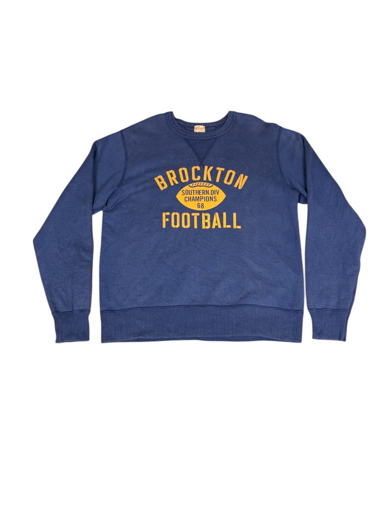 May include: A navy blue sweatshirt with the text "BROCKTON FOOTBALL" in yellow. The text is arched above a yellow football graphic. Below the graphic is the text "SOUTHERN DIV CHAMPIONS 68". The sweatshirt has ribbed cuffs and a crew neck.