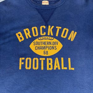 May include: A navy blue sweatshirt with a yellow graphic that reads "BROCKTON FOOTBALL" with a football graphic and the text "SOUTHERN.DIV CHAMPIONS 68". The sweatshirt has a crew neck and a small tag.