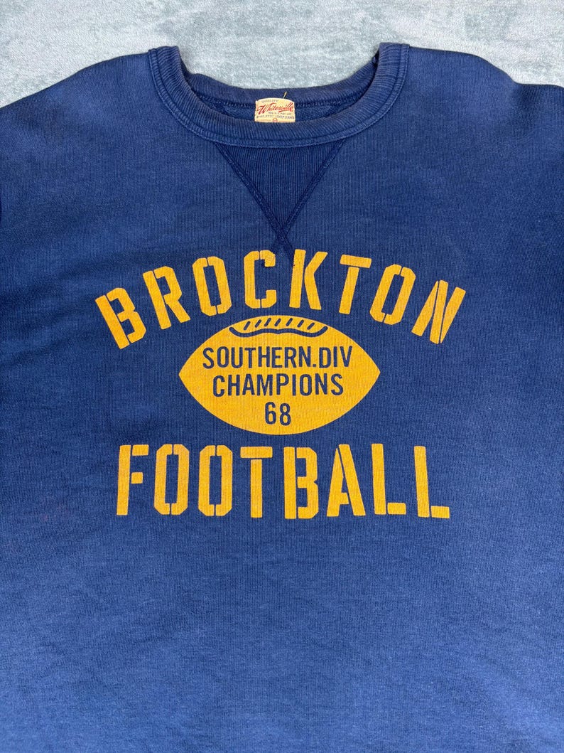 May include: A navy blue sweatshirt with a yellow graphic that reads "BROCKTON FOOTBALL" with a football graphic and the text "SOUTHERN.DIV CHAMPIONS 68". The sweatshirt has a crew neck and a small tag.