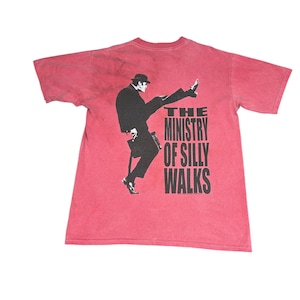 Puede incluir: Camiseta roja con un gráfico en blanco y negro de un hombre con traje haciendo una caminata absurda. El texto "THE MINISTRY OF SILLY WALKS" está impreso debajo del gráfico en una fuente en negrita.