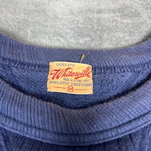 May include: Close-up of a blue sweatshirt with a ribbed crew neck. A rectangular tag with the text "Whitesville" and "Athletic Uniforms" is sewn on the garment. The tag is a light tan color with red and black text.