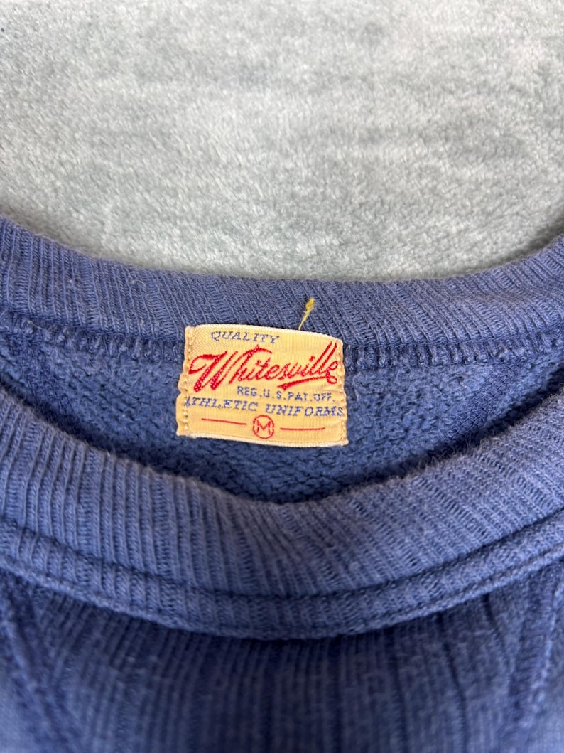 May include: Close-up of a blue sweatshirt with a ribbed crew neck. A rectangular tag with the text "Whitesville" and "Athletic Uniforms" is sewn on the garment. The tag is a light tan color with red and black text.