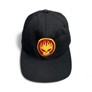 May include: Black baseball cap with a round, embroidered patch featuring a yellow skull with flames and a red border. The cap is made of a solid black fabric and has a curved brim.