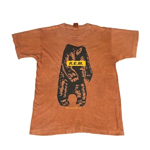 May include: A brown t-shirt featuring a black bear graphic with the text "R.E.M." in a yellow rectangle. The shirt has a vintage look and feel, with a faded appearance.