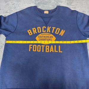 May include: A navy blue sweatshirt with the text "BROCKTON SOUTHERN.DIV CHAMPIONS FOOTBALL" in orange. The sweatshirt has a crew neck and ribbed cuffs and hem. A yellow measuring tape is laid across the front.