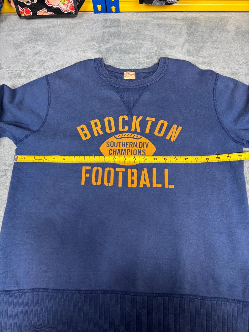 May include: A navy blue sweatshirt with the text "BROCKTON SOUTHERN.DIV CHAMPIONS FOOTBALL" in orange. The sweatshirt has a crew neck and ribbed cuffs and hem. A yellow measuring tape is laid across the front.