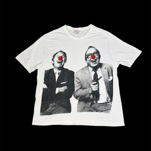 Peut inclure: Un t-shirt blanc à manches courtes avec une impression photographique en noir et blanc représentant deux personnes en costume en train de rire, portant chacune un nez de clown rouge vif. Le motif est centré sur la poitrine du vêtement.