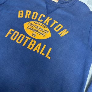 May include: Navy blue sweatshirt with the words "BROCKTON FOOTBALL" in yellow block letters. A yellow football graphic reads "SOUTHERN DIV CHAMPIONS 68". The sweatshirt has a crew neck and a vintage look.