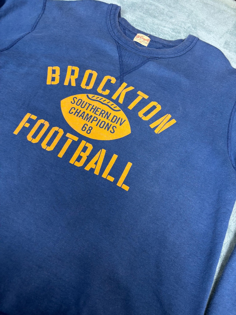 May include: Navy blue sweatshirt with the words "BROCKTON FOOTBALL" in yellow block letters. A yellow football graphic reads "SOUTHERN DIV CHAMPIONS 68". The sweatshirt has a crew neck and a vintage look.