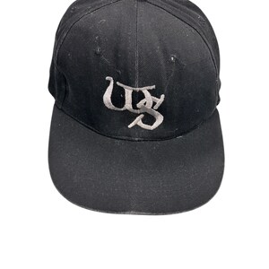 Könnte beinhalten: Schwarze Baseballkappe mit gebogenem Schirm und Knopf oben. Die Kappe hat ein hellgraues gesticktes Design. Das Design scheint die Buchstaben "US" in einer stilisierten Schriftart zu sein.