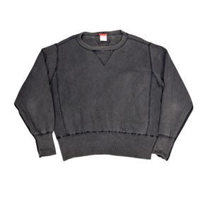 Może przedstawiać: Szara bluza z okrągłym dekoltem i długimi rękawami. Ubranie ma detal w kształcie litery V przy dekolcie oraz prążkowane mankiety i dół. Szwy są widoczne w ciemniejszym odcieniu szarości. Mała czerwona metka jest widoczna.