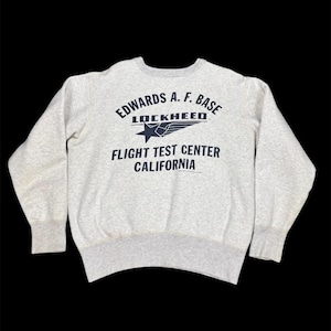May include: A light gray crewneck sweatshirt with the text "EDWARDS A.F. BASE LOCKHEED FLIGHT TEST CENTER CALIFORNIA" in navy blue. The design includes a star and stylized wings. The sweatshirt has ribbed cuffs and hem.