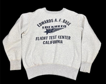 May include: A light grey crew neck sweatshirt featuring navy blue text: "EDWARDS A.F. BASE LOCKHEED FLIGHT TEST CENTER CALIFORNIA". The design includes a star and wings. It has ribbed cuffs and hem.