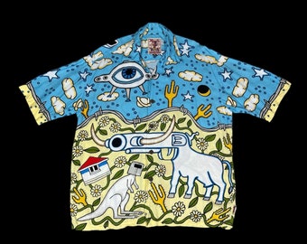 May include: A short-sleeve button-down shirt featuring a light blue upper section with white stars, clouds, and a large eye graphic. The lower section is pale yellow with a white bull-revolver hybrid, a kangaroo with a bucket head, and flowers.