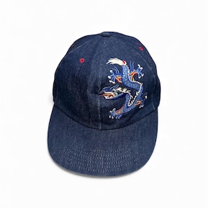 May include: A navy blue denim baseball cap with a curved brim. The cap features an embroidered blue dragon design with white and red accents. The top of the cap has two small red embroidered circles.