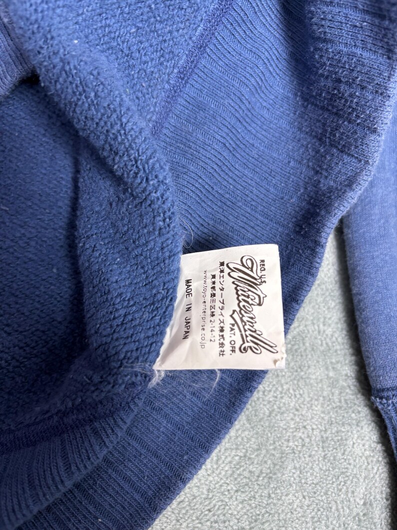 May include: Close-up of a blue sweatshirt with a tag that reads "Wirevale" and "Made in Japan." The tag also includes Japanese text and website information. The sweatshirt has a textured appearance.