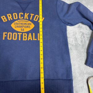 May include: Navy blue sweatshirt with the text "BROCKTON FOOTBALL" in orange block letters. The sweatshirt also features an orange football graphic with the text "SOUTHERN DIV CHAMPIONS 68". The sweatshirt measures approximately 28 inches in length.