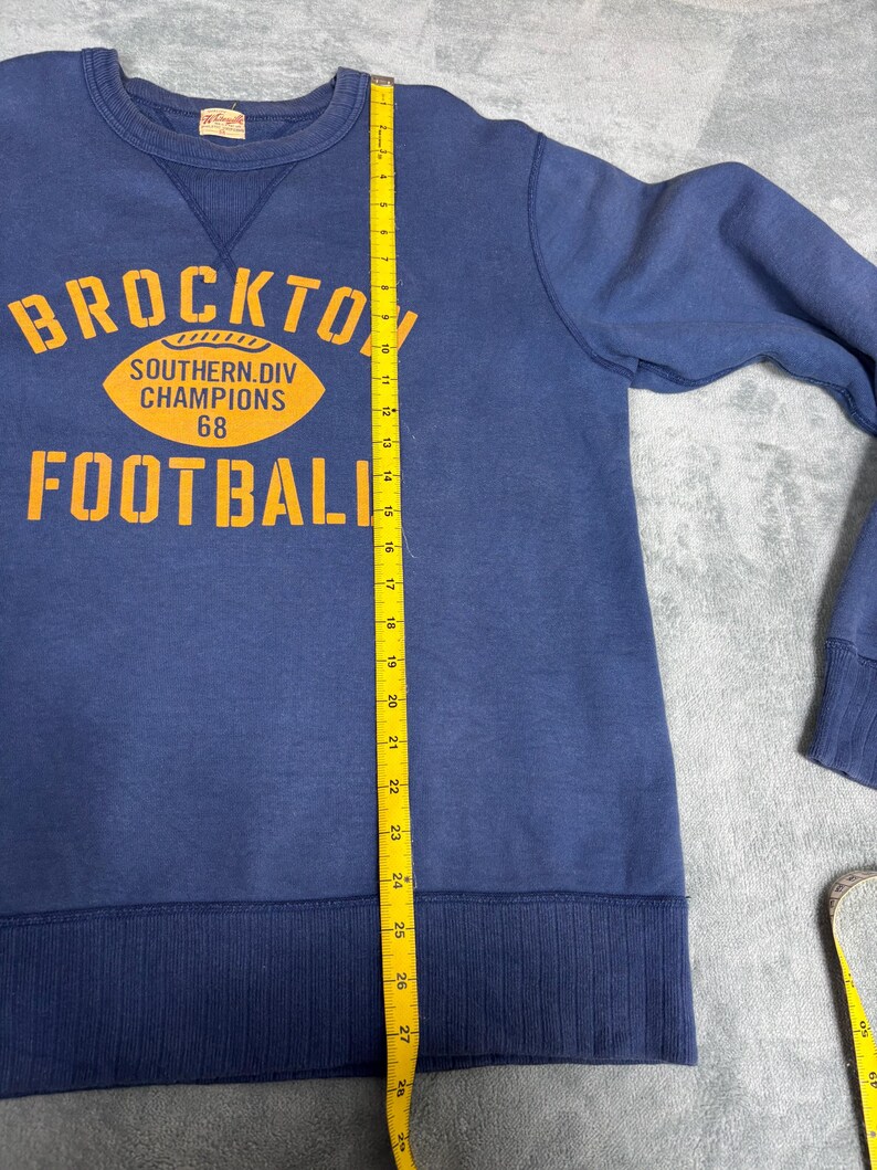 May include: Navy blue sweatshirt with the text "BROCKTON FOOTBALL" in orange block letters. The sweatshirt also features an orange football graphic with the text "SOUTHERN DIV CHAMPIONS 68". The sweatshirt measures approximately 28 inches in length.
