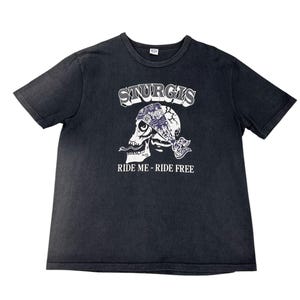 May include: Black t-shirt featuring a skull graphic with a bandana and the word "STURGIS" above it. The text "RIDE ME - RIDE FREE" is below the skull. The shirt has a crew neck and short sleeves.