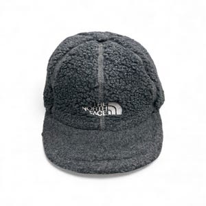 May include: A gray fleece cap with a curved brim. The cap has a textured, fuzzy appearance and features the white "THE NORTH FACE" logo on the front. A vertical seam runs down the center of the cap.