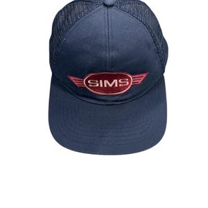 Può includere: Cappellino da baseball blu navy con retro in rete. La parte anteriore del cappellino presenta la scritta "SIMS" in lettere bianche, incorniciata da un ovale e ali rosse. Il cappellino ha una visiera curva e un bottone sulla parte superiore.