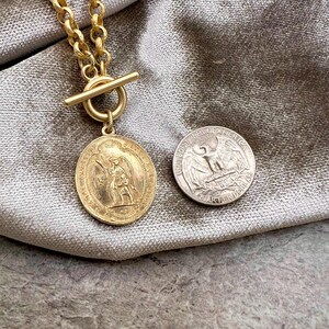 Pu&ograve; includere: Una collana color oro con un ciondolo circolare con un rilievo dettagliato di una figura. La collana ha una chiusura a levetta e una catena spessa. Una moneta da un quarto di dollaro USA &egrave; accanto al ciondolo per il confronto delle dimensioni.