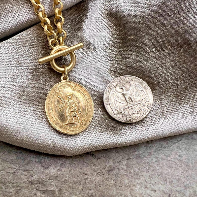 Pu&ograve; includere: Una collana color oro con un ciondolo circolare con un rilievo dettagliato di una figura. La collana ha una chiusura a levetta e una catena spessa. Una moneta da un quarto di dollaro USA &egrave; accanto al ciondolo per il confronto delle dimensioni.