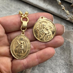 Könnte beinhalten: Zwei goldfarbene religiöse Anhänger. Einer ist oval mit einem Engel-Design, der an einer goldenen Kette hängt. Der andere ist ein runder Anhänger mit einer religiösen Figur. Beide Anhänger haben komplizierte Details und werden vor einem weichen Hintergrund präsentiert.