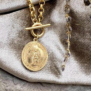 Pu&ograve; includere: Una collana color oro con un ciondolo circolare con un disegno inciso di una figura che tiene un bastone e un'altra figura. La collana ha una chiusura a levetta e una catena con maglie rotonde. Un rametto di lavanda &egrave; visibile sullo sfondo.