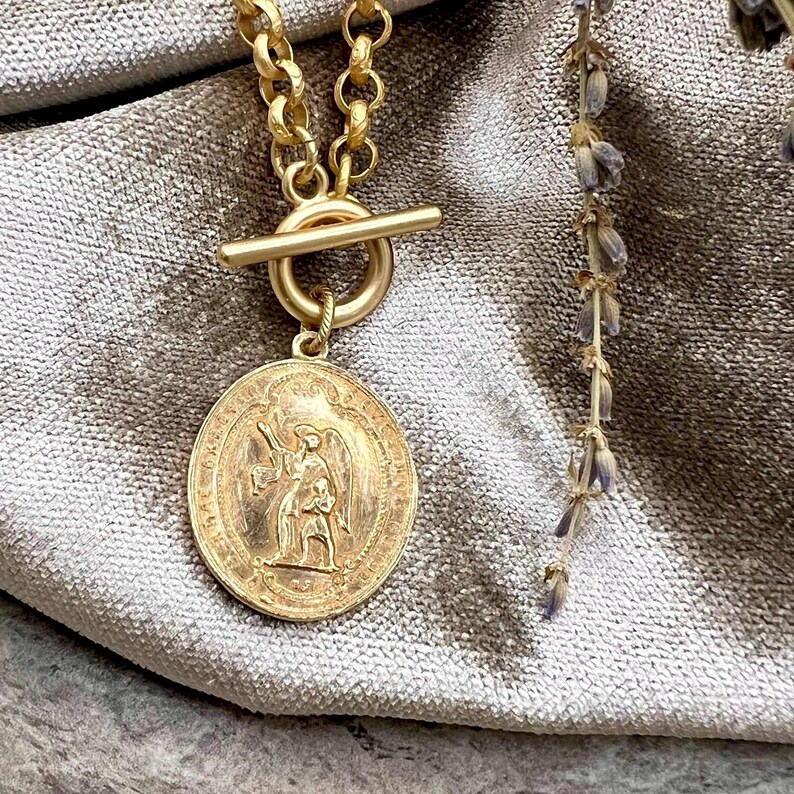 Pu&ograve; includere: Una collana color oro con un ciondolo circolare con un disegno inciso di una figura che tiene un bastone e un'altra figura. La collana ha una chiusura a levetta e una catena con maglie rotonde. Un rametto di lavanda &egrave; visibile sullo sfondo.