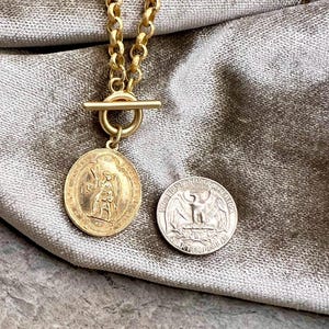 Pu&ograve; includere: Una collana color oro con un grande ciondolo circolare con una figura incisa. La collana ha una chiusura a toggle e una catena con maglie grandi. Una moneta d'argento &egrave; accanto al ciondolo.