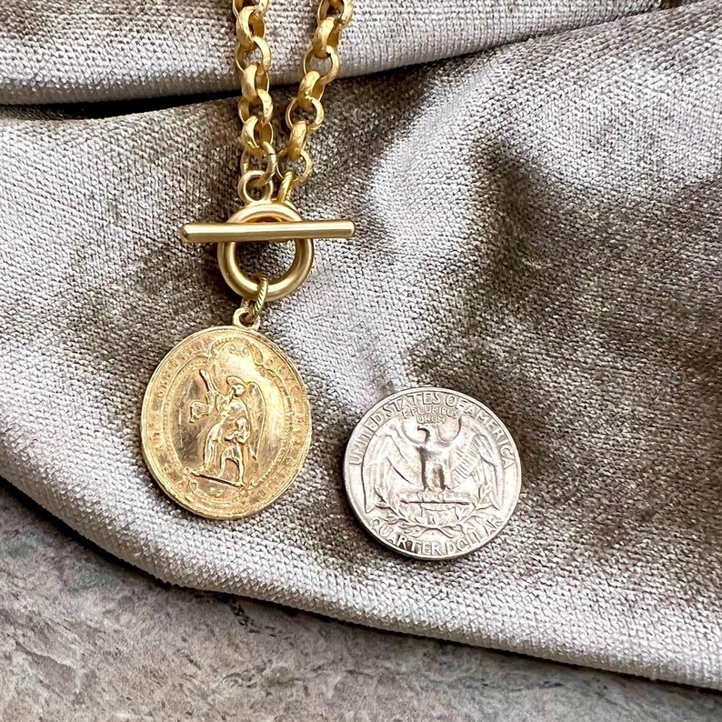 Pu&ograve; includere: Una collana color oro con un grande ciondolo circolare con una figura incisa. La collana ha una chiusura a toggle e una catena con maglie grandi. Una moneta d'argento &egrave; accanto al ciondolo.
