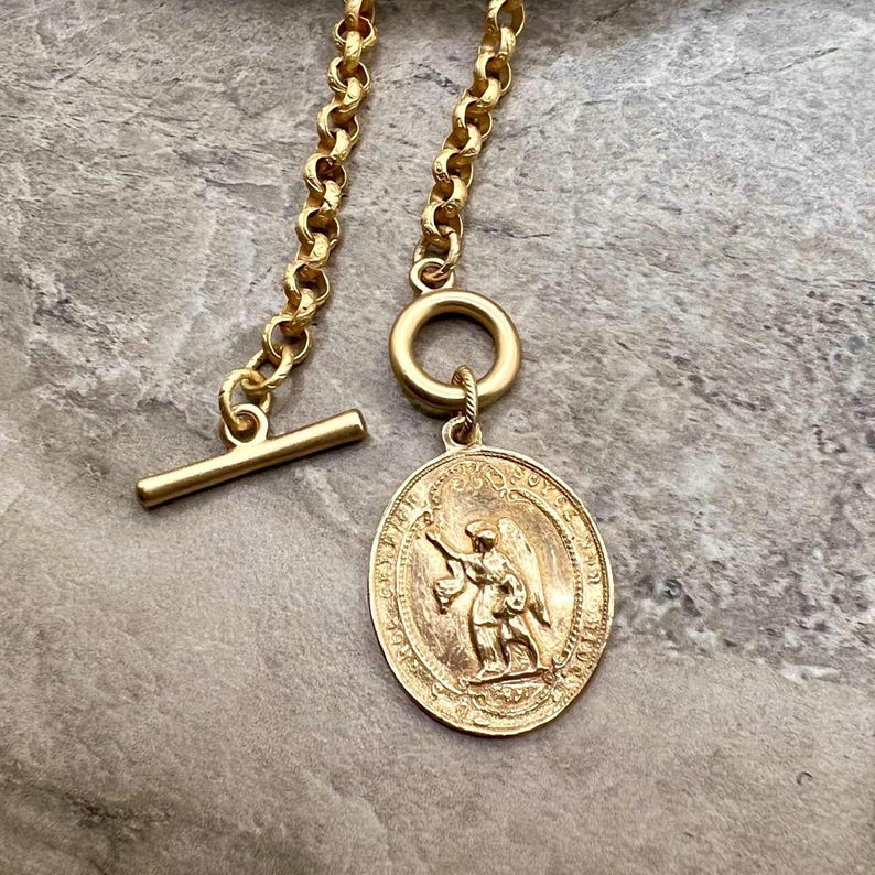 Pu&ograve; includere: Collana dorata con un pendente ovale dettagliato con una figura incisa. La collana ha una chiusura a levetta e una catena con maglie arrotondate. Il pendente raffigura una figura alata che tiene un'arma.