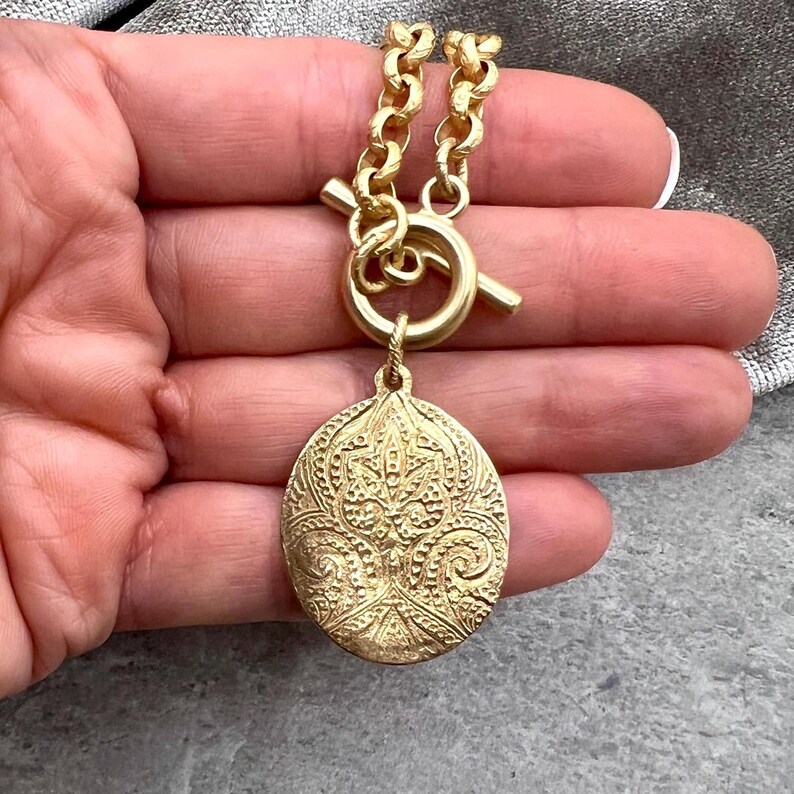 Pu&ograve; includere: Una collana color oro con un pendente ovale dettagliato. Il pendente presenta un design ornato e in rilievo. La collana ha una chiusura a toggle e una catena spessa. Il gioiello &egrave; esposto su uno sfondo neutro.