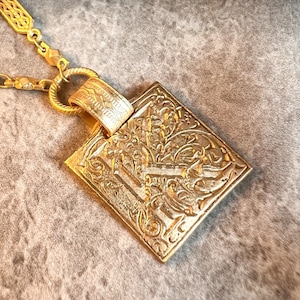 May include: A gold chain necklace with a square pendant featuring an intricate floral design.