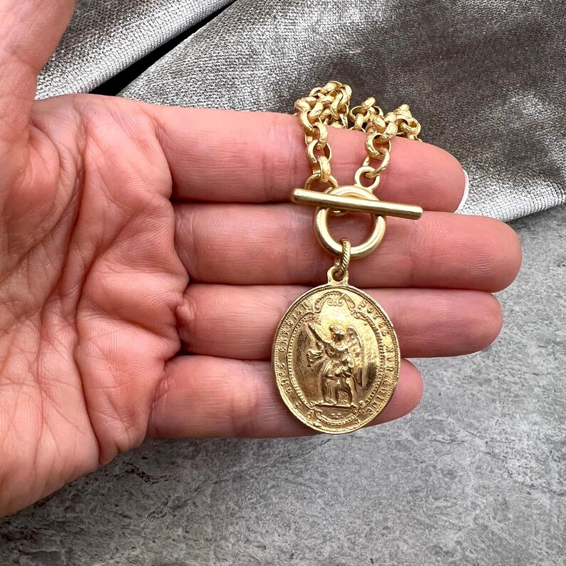 Pu&ograve; includere: Collana dorata con un ciondolo circolare con una figura incisa. La collana ha una catena spessa e una chiusura a levetta. Il ciondolo raffigura un'immagine dettagliata di un santo o un angelo. Il gioiello &egrave; tenuto in una mano.