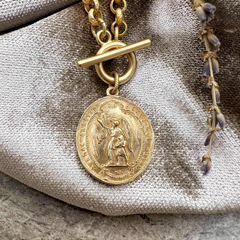 Pu&ograve; includere: Collana color oro con un grande ciondolo circolare con un disegno in rilievo di una figura alata. La collana ha una chiusura a levetta e una catena spessa. Lo sfondo &egrave; un tessuto grigio morbido e si vede un rametto di lavanda.
