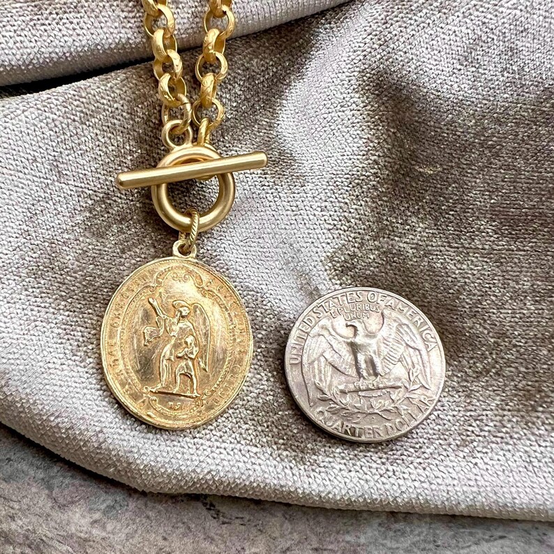 Pu&ograve; includere: Collana color oro con un ciondolo circolare con un rilievo dettagliato di una figura. La collana ha una chiusura a levetta e una catena spessa. Una moneta da un quarto di dollaro USA &egrave; accanto al ciondolo per il confronto delle dimensioni.
