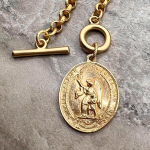 Pu&ograve; includere: Collana dorata con un pendente ovale dettagliato raffigurante un angelo. Il pendente &egrave; attaccato a una catena con una chiusura a levetta. La collana ha un'estetica vintage.