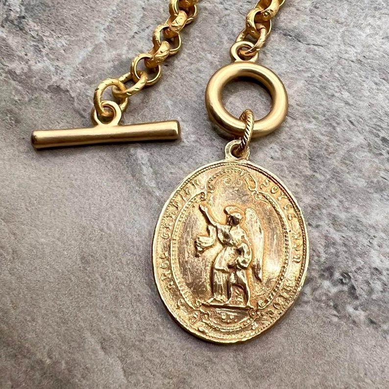Pu&ograve; includere: Collana dorata con un pendente ovale dettagliato raffigurante un angelo. Il pendente &egrave; attaccato a una catena con una chiusura a levetta. La collana ha un'estetica vintage.