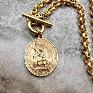Pu&ograve; includere: Collana color oro con un grande ciondolo dettagliato raffigurante un angelo. Il ciondolo &egrave; attaccato a una catena spessa con una chiusura a levetta. La collana &egrave; esposta su una superficie grigia.