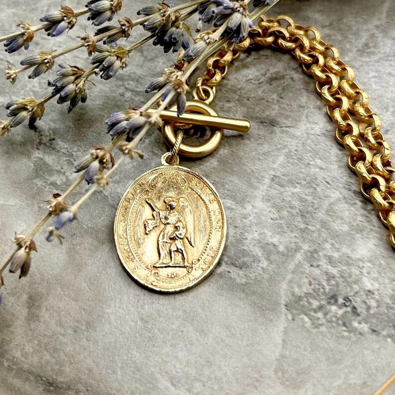 Pu&ograve; includere: Una collana color oro con un ciondolo dettagliato raffigurante una figura di angelo. La collana ha una chiusura a toggle e una catena spessa. Rami di lavanda sono posti vicino alla collana, aggiungendo un tocco di colore naturale.