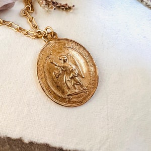 May include: A gold chain necklace with a large, round pendant featuring a detailed embossed image of an angel with wings. The pendant has a raised border and intricate details.